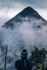 young man hiker contemplating the view from the camp area of the Acatenango Volcano where the Fuego Volcano is seen partially covered with fog on a picturesque afternoon with dense forest