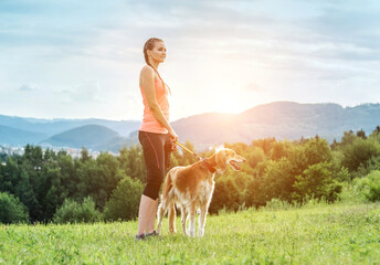 Woman runner and dog on field under golden sunset sky in evening time. Outdoor running. Athletic young man with his dog are running in nature.
