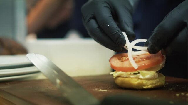 Close up on chef's hands placing tomato and onions on the burger ban. Preparation of the fast-food meal.