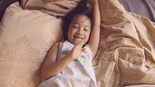 Portrait of adorable asian toddler with white skin in pyjamas who is acting on camera in the bed in the morning with sunlight shining through window. Her eye contact looking expresses joyfulness.