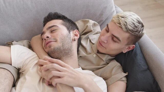 High angle view of young dark-haired Caucasian man and his beloved boyfriend lying together on sofa at home, hugging and talking