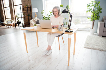 Full length photo of attractive happy nice old woman sit desk look laptop smile indoors inside house home