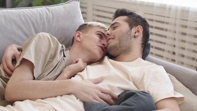Lockdown of young dark-haired Caucasian man and his beloved boyfriend lying together on sofa at home, hugging, talking and kissing