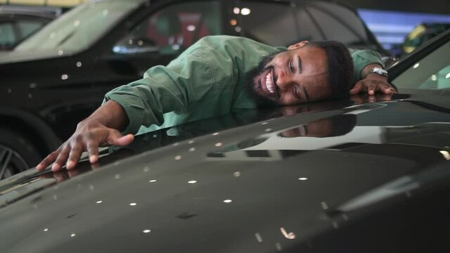 Buy car, happy funny man hugging stroking auto hood that he purchased in dealership. Spbd African