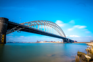 The Bridge at Nijmegen crossing the river Waal and is called there Waalbrug. Long exposure photo on a sunny day with blue sky and a few small clouds.
