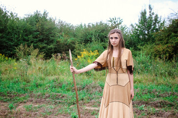 Beautiful young confident woman in american indian dress posing with a spear with the forest in the background, tribal scout concept