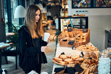 Woman holding cup with coffee looking at pastry, buns, cakes and cookies and waiting for the order. Girl buying a sweet food and hot drink to go. Young woman having a break