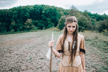 Beautiful young confident woman in american indian dress posing with a spear with the forest in the background, tribal scout concept