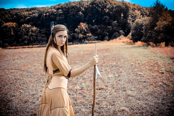 Beautiful young confident woman in american indian dress posing with a spear with the forest in the...