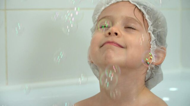 Little smiling girl bathes and plays with soap bubbles in bath. Happy child catches bubbles in white bathroom. Baby bathing. Close up.