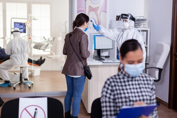 Stomatology receptionist using digital thermometer to measure patient temperature dressed in ppe suit as safety precaution during global outreabk with coronavirus. Dentist treating teeth.