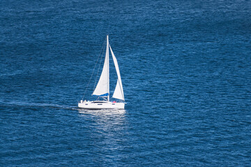 aerial view of a generic white sailboat with two sails sailing on very calm water in the San Diego Bay in Southern California surrounded by small ripples and swells