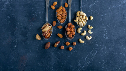 Assortment of various types of nuts: cashew, hazelnuts, almonds, brazil nuts on metal silver spoons on dark background. Healthy vegetarian snacks. Protein-containing food. Copy space for text