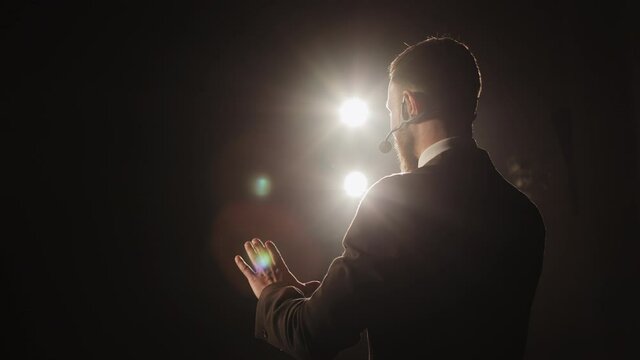 The speaker is conducting the conference. He is standing on the stage and talking into the microphone. He is waving his hands. The spotlight is shining on him. The camera is rotating around him. 4K