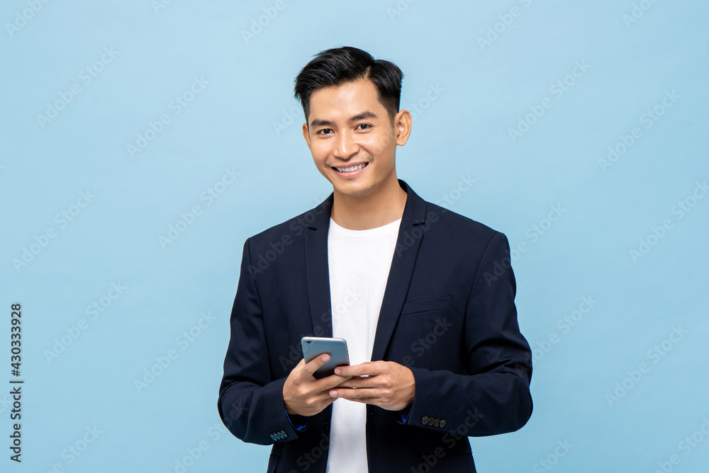 Wall mural young smiling handsome asian man in semi formal suit using mobile phone in light blue isolated studi