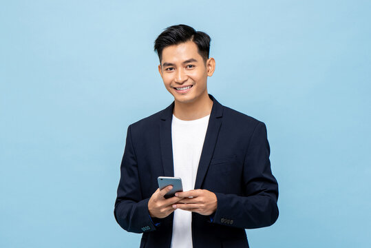 Young smiling handsome Asian man in semi formal suit using mobile phone in light blue isolated studio background