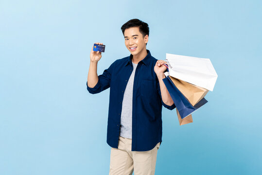 Happy Smiling Handsome Asian Man Carrying Shopping Bags And Showing Credit Card In Light Blue Isolated Studio Background