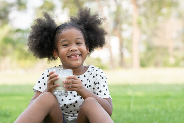 Little African kid girl smiling with milk mess on lips mouth and broken tooth. Cute child holding a glass of milk and sitting on green grass at park on summer