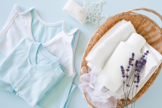 Baby Bodysuits On Light Blue Table Background. Pastel Color. White Bottle On Towel With Dried Lavender Flower In Basket. Care About Clean And Soft Hands, Face, Legs And Body Kid Skin. Top Down View.