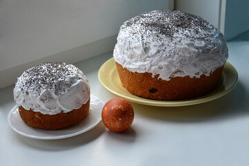 A brown speckled chicken egg and two Easter cakes with white icing and poppy sprinkles on a white plate. Photo on the windowsill. Home cooking for Easter.