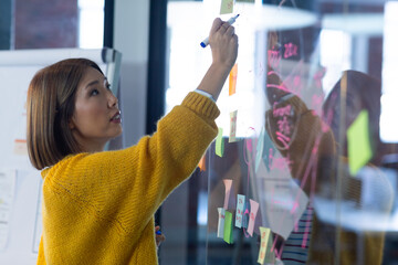 Asian businesswoman standing in front of glass wall and writing in office