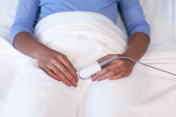 Midsection of mixed race female patient lying in hospital bed wearing fingertip pulse oximeter