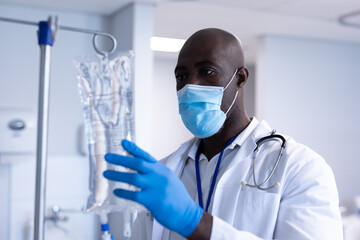 African american male doctor wearing face mask and gloves preparing iv drip bag in hospital ward