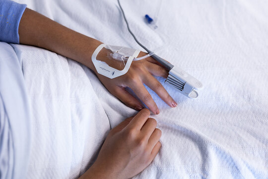 Midsection of mixed race female patient in hospital bed wearing fingertip pulse oximeter and iv tube