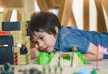 Portrait of School boy lying down on the carpet floor playing with soldiers, military car and figurine toys, Happy Kid playing wars and peace on his own at home, Children imagination and development
