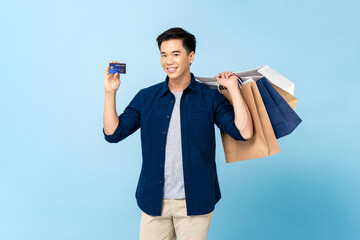 Happy smiling handsome Asian man carrying shopping bags and showing credit card in light blue...