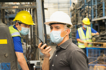 Men work together, wear safety facemask, use walkie-talkie, look straight. Caucasian engineer men  wear safety facemask, use walkie-talkie, look straight while Asian men working behind him in factory