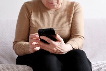 Elderly woman with smartphone sitting on sofa at home, mobile phone in female hands. Concept of online communication in retirement, sms, social media