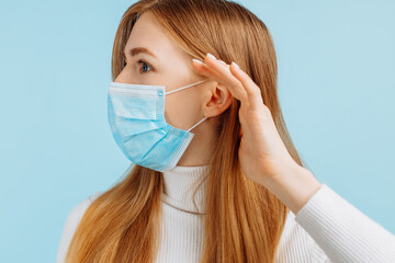 Young woman in a medical mask, on an blue background, listening to something with her hand on her ear