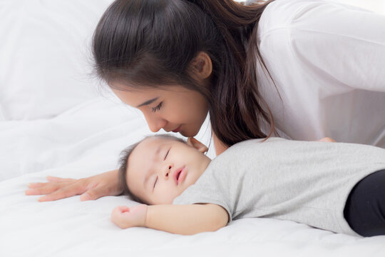 Young asian mother kiss cheek of little baby girl with tender on bed in the bedroom, mom love newborn and care, mother with expression with child together, parent and daughter, family concept.