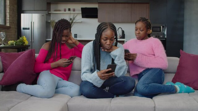 Addicted to cellphone lovely african american teenage girls sitting on couch, phubbing each other, browsing social media content online on smart phones, showing ignorance and apathy indoors.