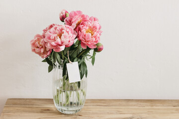 Floral still life scene. Pink peonies flowers, bouquet in glass vase on wooden table. Blank gift...