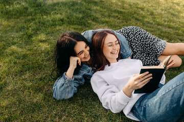 Two best friends spending time outdoors, lying on green grass in a park and reading a book,...