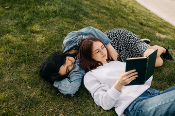 Two sisters lying on green grass in a park and reading a book.
