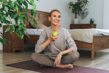 Beautiful blonde young woman in comfortable sport wear practice yoga hold green apple in hand in...
