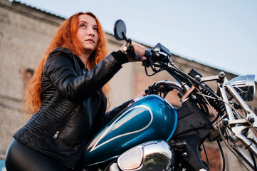 Naklejka na ściany i meble Curly red-haired woman in a black leather jacket sits on a motorcycle. Portrait of a serious girl driving a bike.