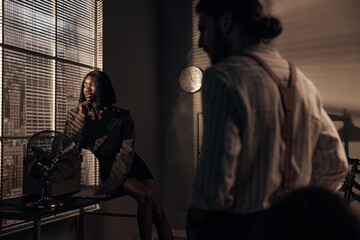 beautiful dark-skinned girl in a gray short jacket sitting on the table and looking out the window overlooking the big city and nearby is a young man in a shirt with suspenders