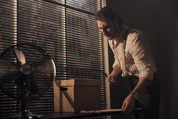 scene of a brutal young detective standing at the table and looking at documents in his office, he is wearing a striped shirt, classic pants and suspenders