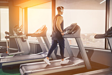 Fototapeta na wymiar Concentrated fit man listening to music in headphones while running on treadmill in modern gym with panoramic windows.