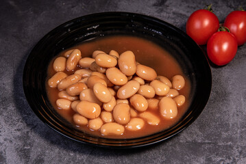 White falo in tomato sauce. Boiled beans, stewed with tomato sauce in a black plate on a dark surface and cherry tomatoes. Close-up