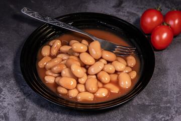 White falo in tomato sauce. Boiled beans, stewed with tomato sauce in a black plate on a dark surface and cherry tomatoes. Close-up