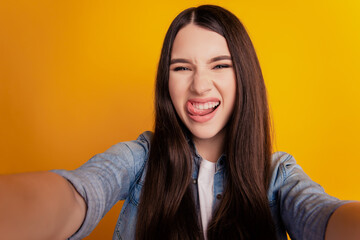 Fototapeta premium Close-up of young crazy funky woman taking selfie stick-out tongue on yellow background