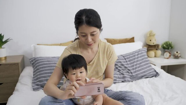 smiling asian mother holding a mobile phone is sitting with her young kid and waiting for friend to answer the phone while making a video call in the bedroom.