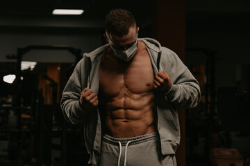 Fototapeta na wymiar A bodybuilder in a face mask to avoid the spread of coronavirus is opening his zipped hoodie to demonstrate his athletic physique. A sporty guy in a surgical mask is posing after a workout in a gym.