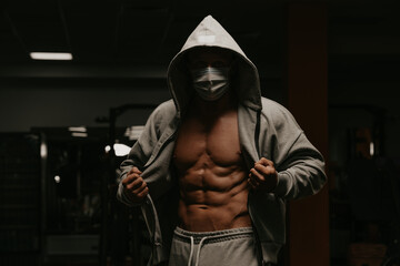 Fototapeta na wymiar A bodybuilder in the hood and a face mask to avoid the spread of coronavirus is opening his zipped hoodie to demonstrate his abs. A sporty guy in a surgical mask is posing after a workout in a gym.