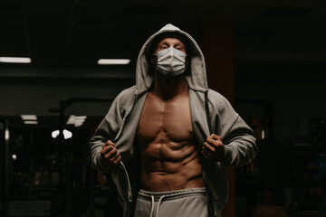 Naklejka na ściany i meble A bodybuilder in a hood and a face mask to avoid the spread of coronavirus is opening a hoodie to demonstrate his athletic physique. A sporty guy in a surgical mask is posing after a workout in a gym.